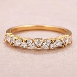 Puede incluir: Un anillo de oro con piedras preciosas claras. El anillo presenta un patrón repetido de piedras redondas y en forma de marquesa, engastadas en una delicada banda de oro amarillo. El anillo se muestra sobre un fondo rosa suave.