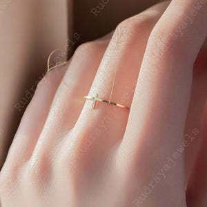 Pode incluir: Um anel de ouro simples com um pequeno design de cruz. O anel &eacute; usado em um dedo.