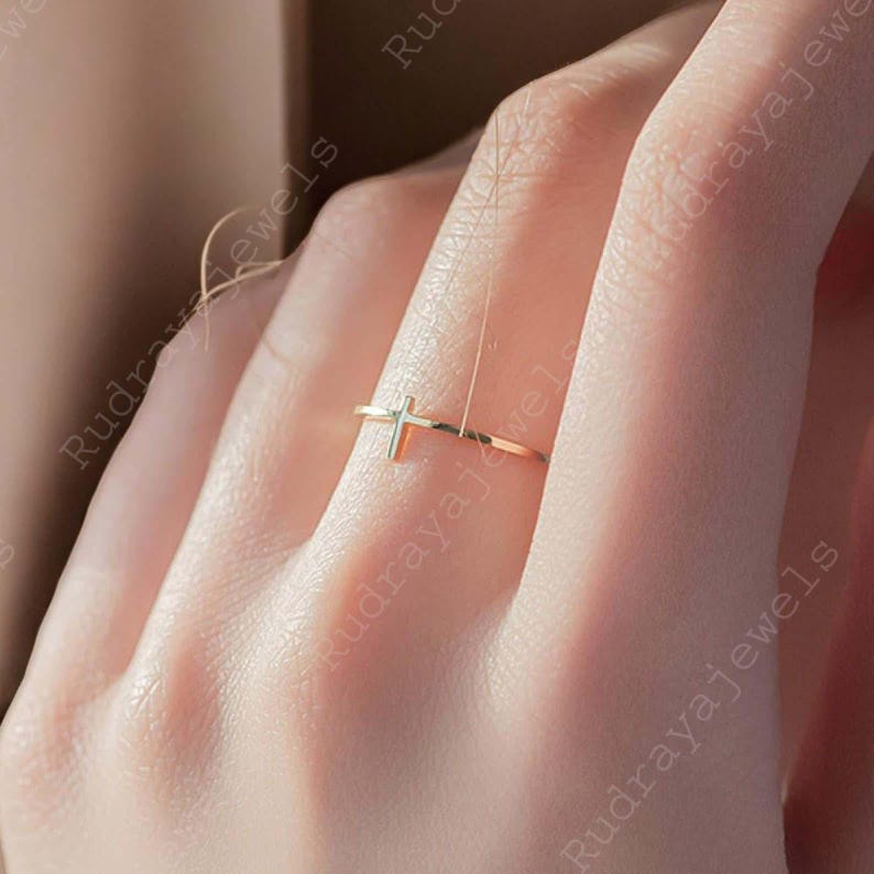 Pode incluir: Um anel de ouro simples com um pequeno design de cruz. O anel &eacute; usado em um dedo.