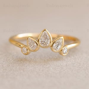 Puede incluir: Un anillo de oro con diseño de corona, con tres diamantes en forma de pera y dos diamantes redondos más pequeños. El anillo tiene un acabado pulido. El fondo es neutro.