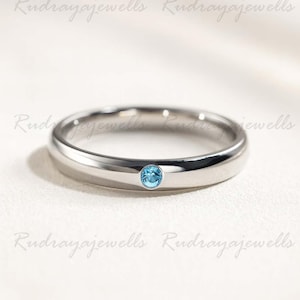 Puede incluir: Un anillo de banda de metal en tono plateado pulido que presenta una &uacute;nica piedra preciosa peque&ntilde;a, redonda y de color azul claro, engastada al ras en el centro de la banda. El anillo tiene un perfil liso y redondeado.