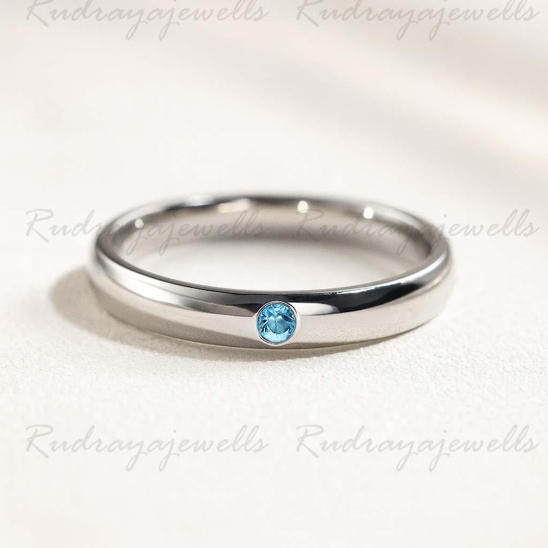 Puede incluir: Un anillo de banda de metal en tono plateado pulido que presenta una &uacute;nica piedra preciosa peque&ntilde;a, redonda y de color azul claro, engastada al ras en el centro de la banda. El anillo tiene un perfil liso y redondeado.