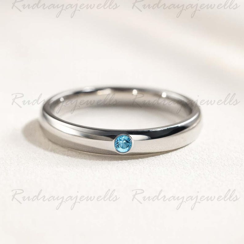 Puede incluir: Un anillo de banda de metal en tono plateado pulido que presenta una &uacute;nica piedra preciosa peque&ntilde;a, redonda y de color azul claro, engastada al ras en el centro de la banda. El anillo tiene un perfil liso y redondeado.