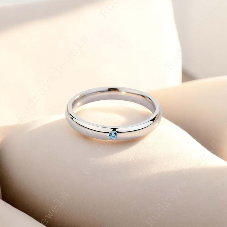Puede incluir: Un anillo de banda de metal en tono plateado pulido que presenta una &uacute;nica piedra preciosa peque&ntilde;a, redonda y de color azul claro, engastada al ras en el centro de la banda. El anillo tiene un perfil liso y redondeado con una superficie brillante.