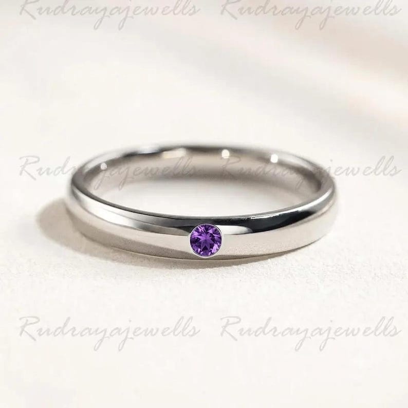 Puede incluir: Un anillo de banda de metal en tono plateado pulido que presenta una &uacute;nica piedra preciosa de amatista morada de corte redondo, engastada al ras en el centro de la banda. El anillo tiene un perfil redondeado liso y un acabado reflectante.