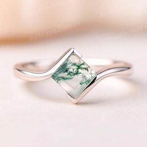 May include: A silver ring featuring a square-cut, moss agate gemstone with green inclusions. The ring has a delicate, curved band and a modern, minimalist design. The gemstone is set in a bezel setting.