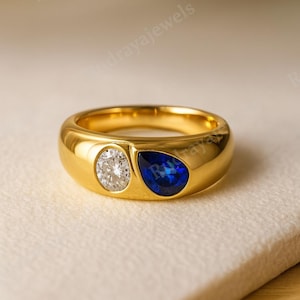 May include: A gold ring featuring a round, clear diamond and a teardrop-shaped, deep blue gemstone. The ring has a thick, polished band and is set on a neutral surface. The ring is a piece of jewelry.