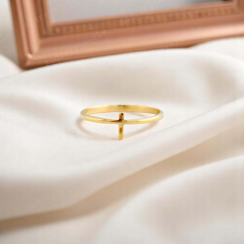 Pode incluir: Um anel dourado com um design de cruz simples. O anel est&aacute; sobre um fundo de tecido branco.