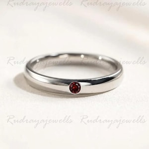 Puede incluir: Un anillo de banda de metal en tono plateado pulido que presenta una &uacute;nica piedra preciosa granate peque&ntilde;a, redonda y de color rojo intenso, engastada al ras en el centro de la banda. El anillo tiene un perfil liso y redondeado.