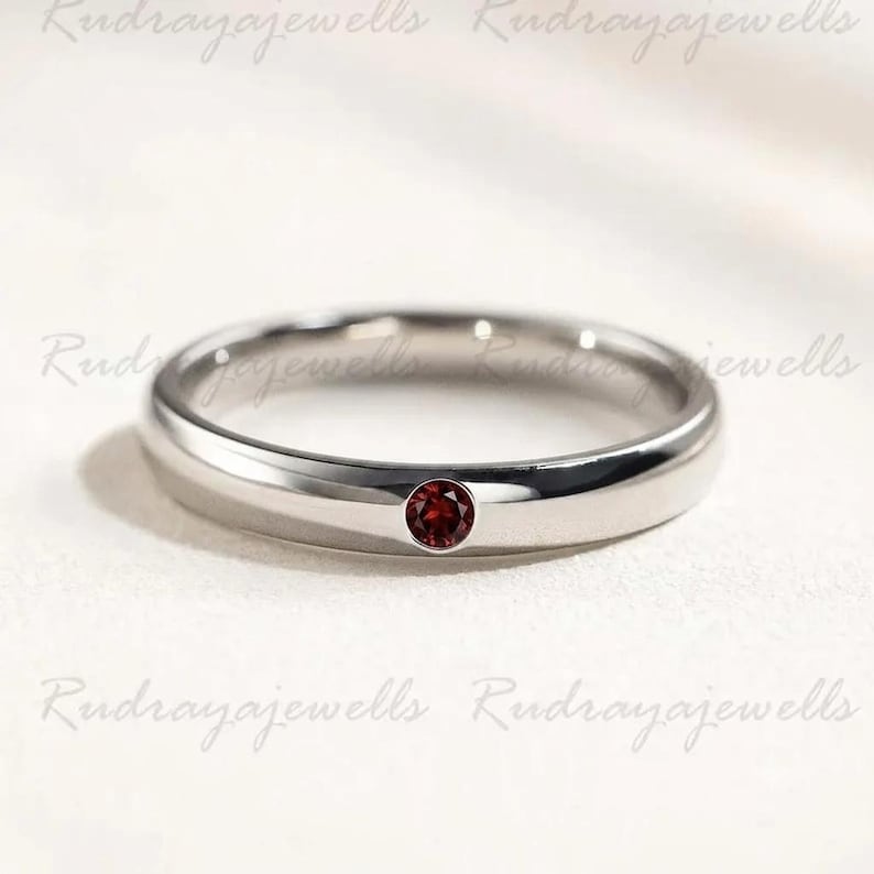 Puede incluir: Un anillo de banda de metal en tono plateado pulido que presenta una &uacute;nica piedra preciosa granate peque&ntilde;a, redonda y de color rojo intenso, engastada al ras en el centro de la banda. El anillo tiene un perfil liso y redondeado.