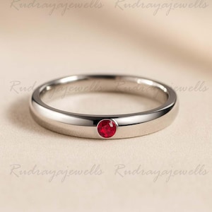 Könnte beinhalten: Ein polierter Silberring mit einem einzelnen, runden, leuchtend roten Edelstein. Der Ring hat ein glattes, abgerundetes Band und ein minimalistisches Design. Der Edelstein ist in einer einfachen Lünettenfassung gefasst.