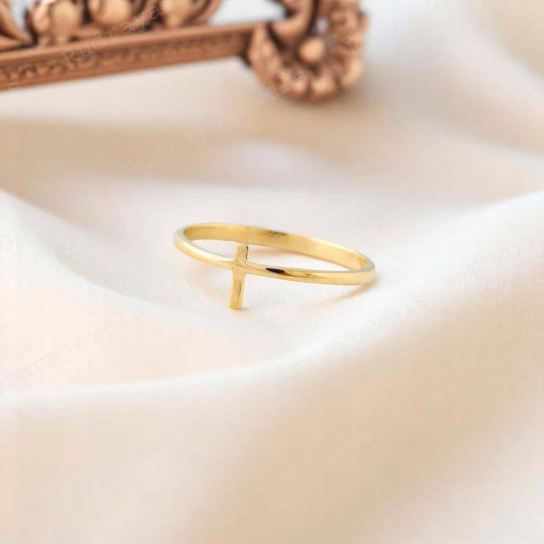 Pode incluir: Um anel dourado com um design de cruz simples. O anel &eacute; fino e delicado, e a cruz est&aacute; centralizada na banda.