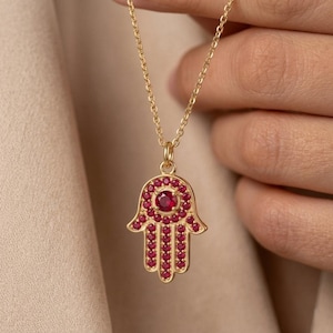 May include: A gold-colored necklace featuring a Hamsa hand pendant. The pendant is adorned with numerous small, red gemstones and a larger, central red gemstone. The necklace is suspended from a delicate gold chain.