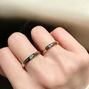 Puede incluir: Dos anillos de metal liso en dedos adyacentes, cada uno con una peque&ntilde;a piedra preciosa redonda de color azul claro engastada al ras en el centro de la banda. Un anillo es de tono plateado y el otro es de tono oro rosa.