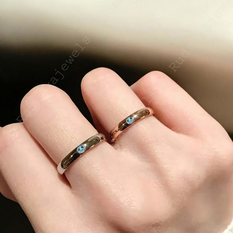 Puede incluir: Dos anillos de metal liso en dedos adyacentes, cada uno con una peque&ntilde;a piedra preciosa redonda de color azul claro engastada al ras en el centro de la banda. Un anillo es de tono plateado y el otro es de tono oro rosa.