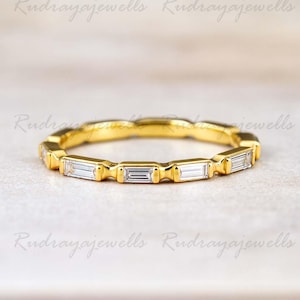 May include: A gold band ring featuring rectangular-cut gemstones. The ring has a continuous design of alternating gold segments and clear, rectangular stones. The ring is set against a soft, neutral background.