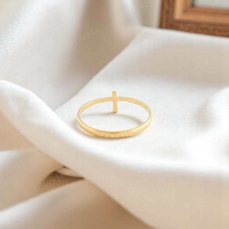 Pode incluir: Um anel dourado com um pequeno design de cruz. O anel est&aacute; sobre um fundo de tecido branco.