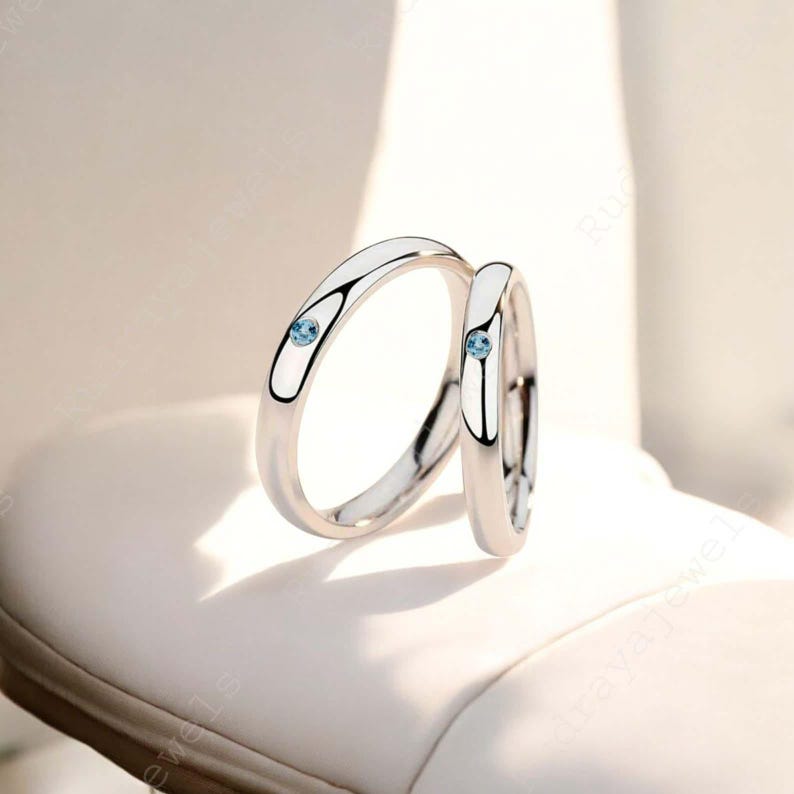 Puede incluir: Dos anillos de metal en tono plateado pulido, cada uno con una peque&ntilde;a piedra preciosa redonda de color azul claro engastada al ras en la banda. Los anillos tienen un perfil liso y redondeado con un acabado reflectante y un dise&ntilde;o limpio.