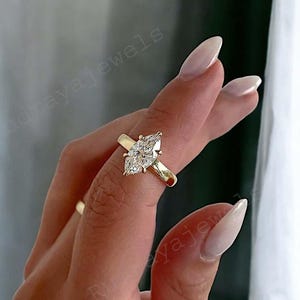 Peut inclure: Une bague de fiançailles en or avec un diamant taille marquise. La bague a un anneau simple et une monture à griffes qui met en valeur la forme du diamant. La bague est portée au doigt.