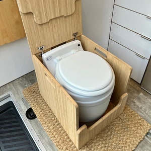 May include: A white toilet bowl with a wooden lid and frame. The toilet is in a small bathroom with a sink and cabinets.