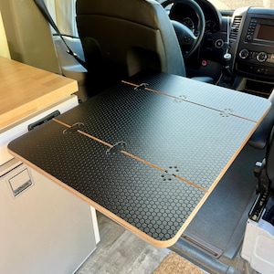 May include: A black folding table with a honeycomb pattern surface is mounted to the wall of a van. The table is folded up and is in the closed position. The table is made of wood and has a black finish.