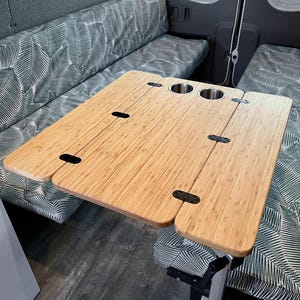May include: A folding bamboo table with two cup holders. The table is made of natural bamboo and has a light brown color. The table is folded in half and is sitting on a gray patterned fabric.