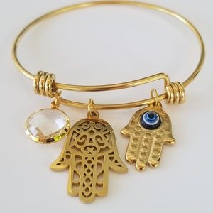 Puede incluir: Pulsera de oro con tres dijes: un cristal transparente, una mano de hamsa con intrincados recortes y una mano de hamsa con un ojo turco azul.
