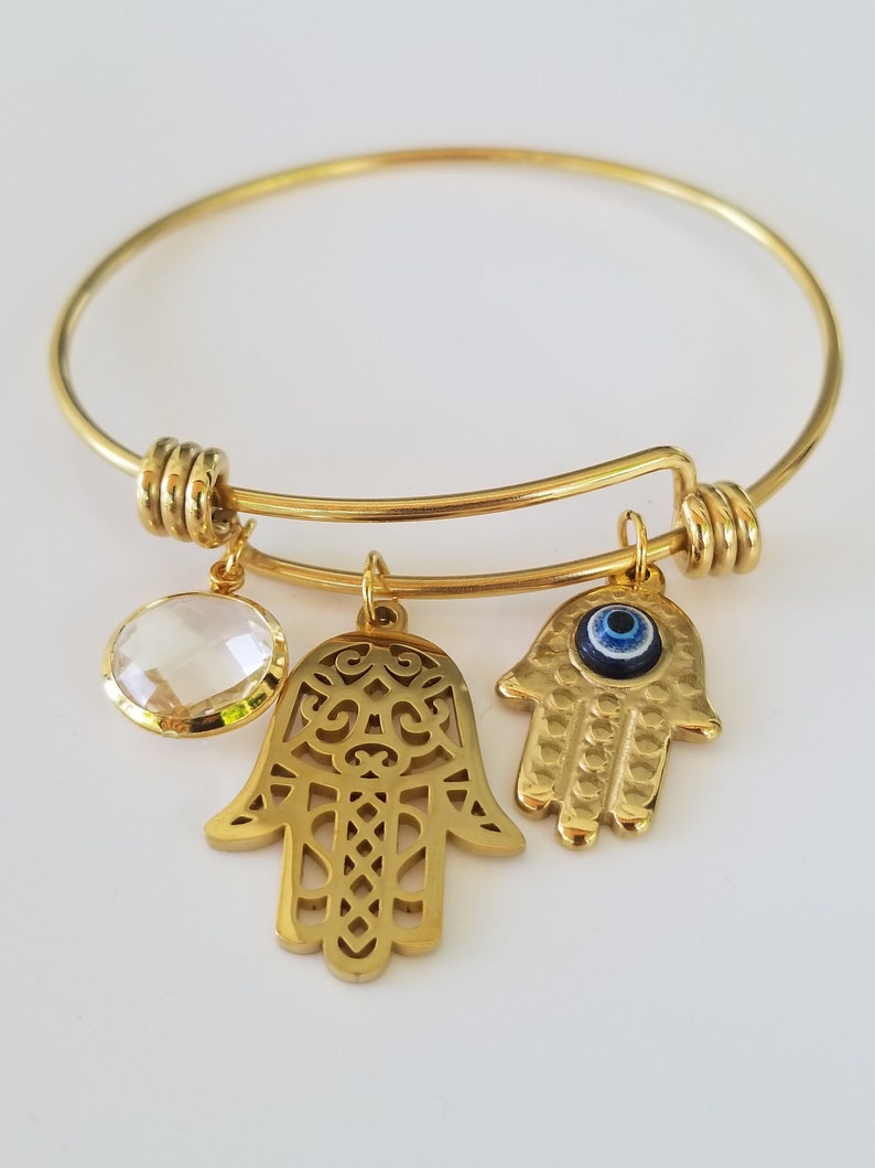 Puede incluir: Pulsera de oro con tres dijes: un cristal transparente, una mano de hamsa con intrincados recortes y una mano de hamsa con un ojo turco azul.
