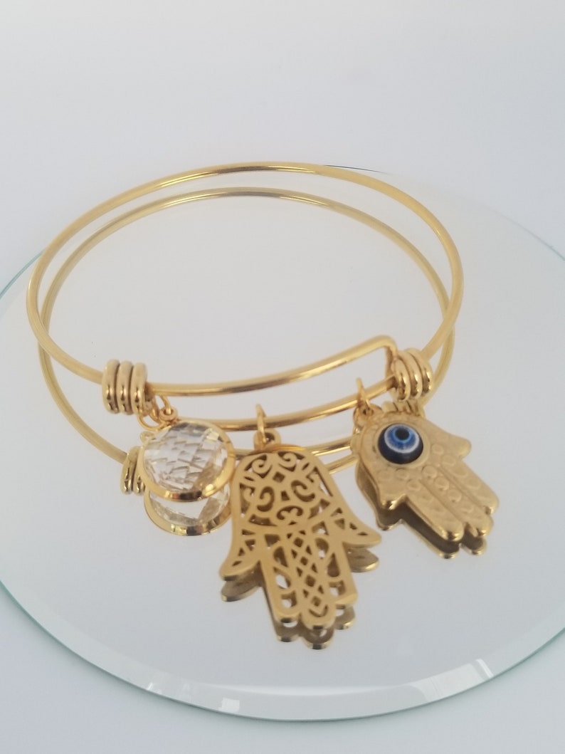 Puede incluir: Pulsera de oro con tres dijes: un cristal transparente, una mano de hamsa y una mano de hamsa con un ojo turco azul.