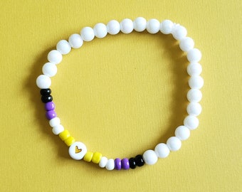 Könnte beinhalten: Ein weißes Armband mit einem Herz-Anhänger in der Mitte. Der Herz-Anhänger ist gelb, lila und schwarz und repräsentiert die Non-Binary-Pride-Flagge.