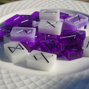 May include: A pile of purple and white rune stones with silver glitter. The runes are carved into the stones and are visible on both sides.