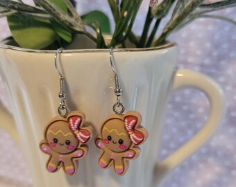 May include: A pair of silver dangle earrings featuring gingerbread person charms with pink and white icing details. The gingerbread people have smiling faces and pink bows.
