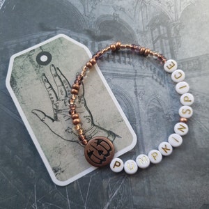May include: A white beaded bracelet with gold lettering spelling "Pumpkin Spice". The bracelet has a copper charm in the shape of a pumpkin.
