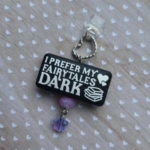 Puede incluir: Un dije rectangular negro con el texto "I PREFER MY FAIRYTALES DARK" en blanco. El dije tiene un corazón y un icono de libro. Un anillo plateado en forma de corazón se conecta a un clip transparente. Una estrella y una cuenta moradas cuelgan debajo.