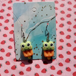 May include: A pair of dangle earrings featuring green frog popsicle charms with orange and white accents. The earrings are displayed on a blue and white card with a white circle in the top corner.