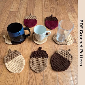 May include: Six crocheted acorn-shaped coasters in various colors, including brown, tan, and red. The coasters are placed on a wooden surface.
