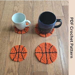 Puede incluir: Cuatro posavasos de baloncesto de ganchillo naranja y negro con dos tazas. Los posavasos son redondos con líneas negras que imitan el diseño de una pelota de baloncesto. El texto "PDF Crochet Pattern" es visible.