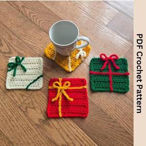 May include: A collection of four handmade crochet coasters in the shape of gift boxes. The coasters are in various colors, including red, green, yellow, and cream, with contrasting ribbon accents. A white mug sits on a yellow coaster. The image includes the text "PDF Crochet Pattern".