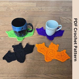 May include: Four crocheted bat-shaped coasters in green, purple, black, and orange. The coasters are placed on a wooden floor with two mugs on top of two of the coasters.