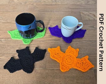 May include: Four crocheted bat-shaped coasters in green, purple, black, and orange. The coasters are placed on a wooden floor with two mugs on top of two of the coasters.
