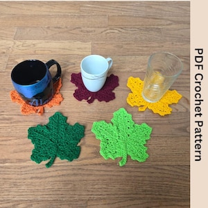 Puede incluir: Cinco posavasos de ganchillo en forma de hoja en diferentes colores: naranja, granate, amarillo, verde y verde lima. Los posavasos están sobre una superficie de madera con dos tazas y un vaso encima.