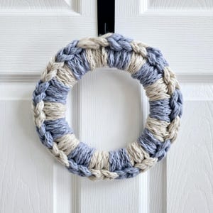 May include: A blue and beige crocheted wreath hanging on a white door. The wreath is made of thick rope and has a braided design.
