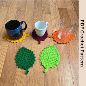 May include: Four crocheted leaf-shaped coasters in green, orange, yellow, and maroon. The coasters are on a wooden surface and are being used to hold mugs and a glass.