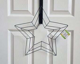 May include: Black metal star-shaped wreath form hanging on a white door. The star is made of wire and has a layered design. A black over-the-door hanger is used to display the wreath. A small tag is attached to the star.