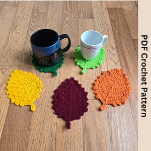 Puede incluir: Cuatro posavasos de ganchillo en forma de hoja en amarillo, naranja, verde y burdeos. Los posavasos están sobre una superficie de madera con dos tazas.
