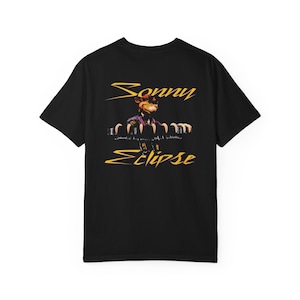 May include: Black t-shirt featuring a graphic of a bear wearing sunglasses and a purple suit. The text "Sonny Eclipse" is written in yellow cursive above and below the bear illustration. The shirt is made of a soft, comfortable material.