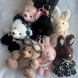 May include: A collection of plush toys, including a brown teddy bear wearing goggles, a pink bunny with a pearl necklace, a white bunny with a pearl necklace, a brown bunny with a black and white dress, a white bear with a black and blue outfit, and a black poodle.