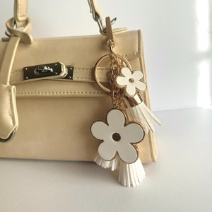 May include: A cream-coloured handbag with a gold-tone chain and a white flower charm with a tassel.