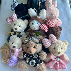 May include: A collection of plush teddy bear keychains with various accessories, including a pink bunny, a black and white checkered bow, a brown bear with a red and white checkered bow, a white bear with a brown hat, a brown bear with a camera, and a pink bear with a pink bow.