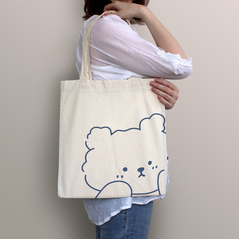 May include: A white canvas tote bag with a blue outline of a cartoon bear's head. The bear is looking to the left and has a fluffy appearance.